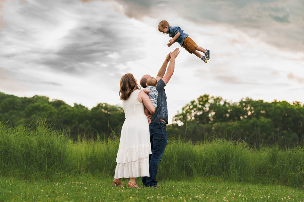 A family in a tall grassy field are tossing their toddler in the air with a dramatic sky behind them.