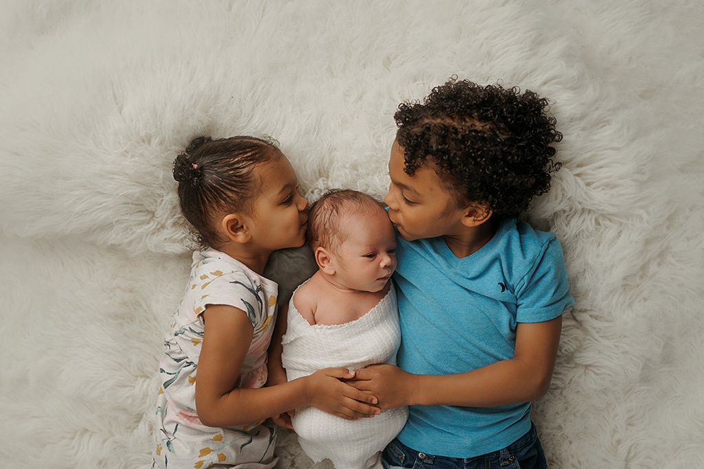 Siblings are giving their newborn brother a kiss on his head while snuggling on a fur blanket.