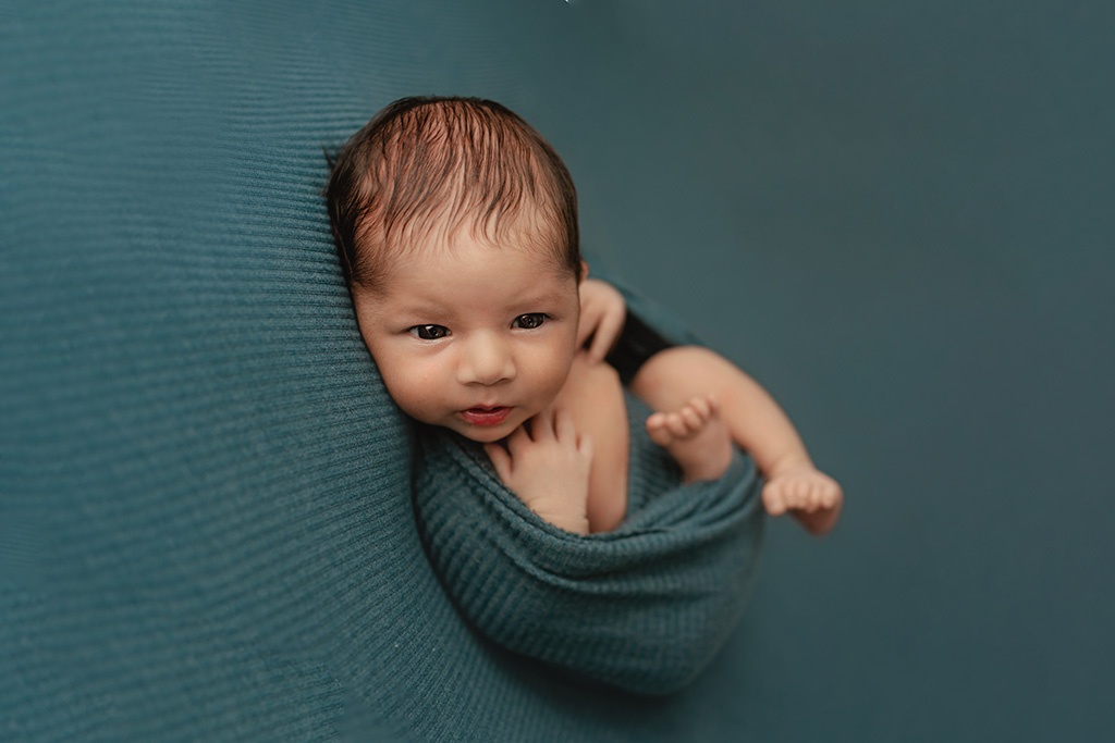A newborn boy is wrapped in a blue swaddle looking at the camera during his newborn photo session.