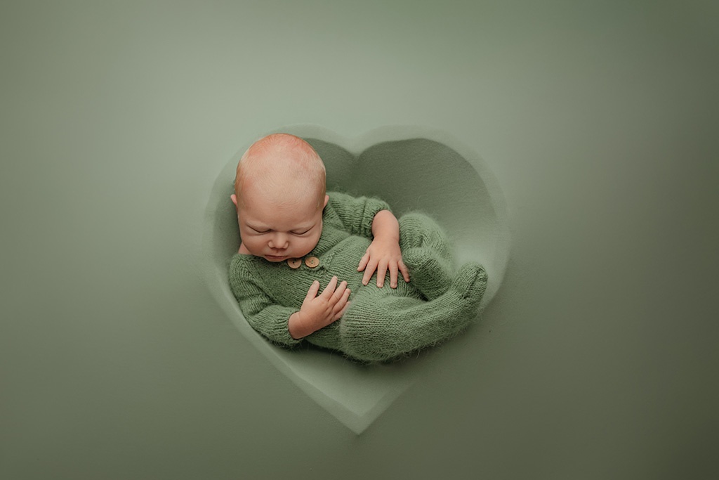 An infant boy in a knit onesie in laying on a green heart shaped bowl during his newborn photography session in Owatonna, MN.